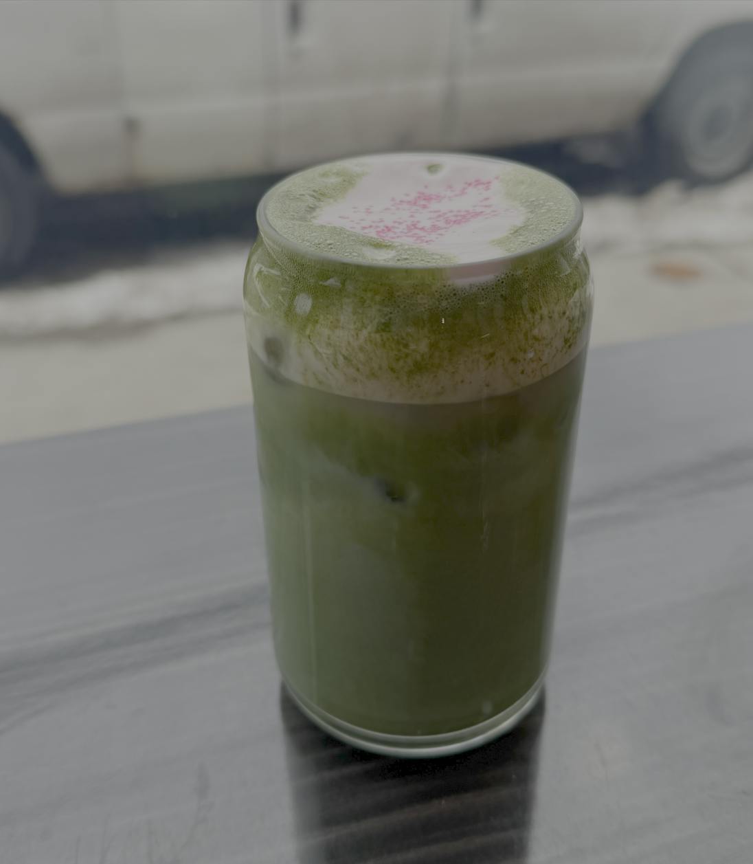 An iced, green matcha latte sits on a wooden table with pink cream on top. 