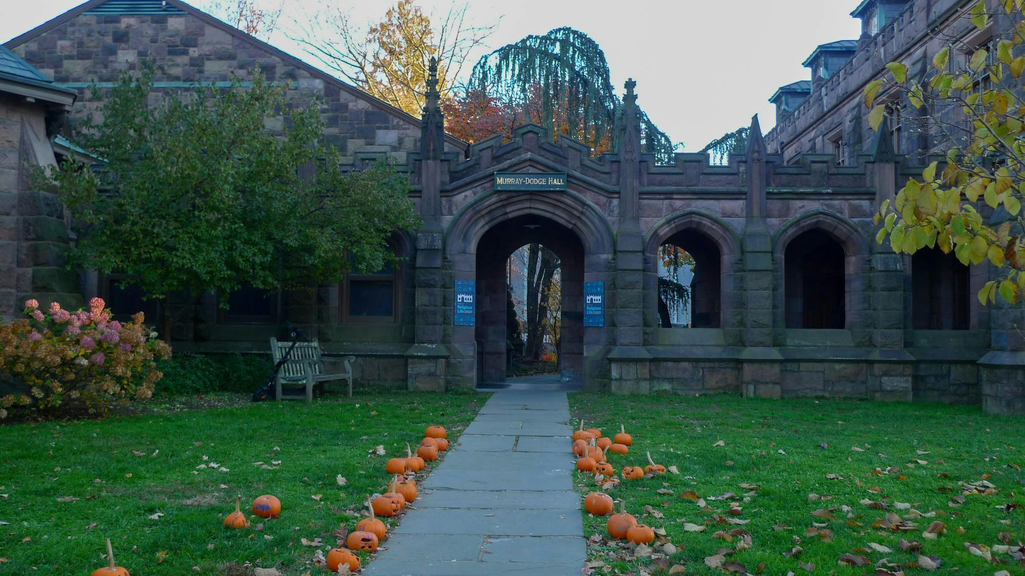 Pumpkins line a stone path to a brown stone building. There are trees and bushes framing the building.