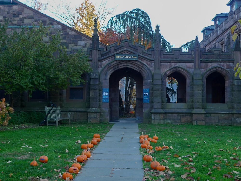 Pumpkins line a stone path to a brown stone building. There are trees and bushes framing the building.
