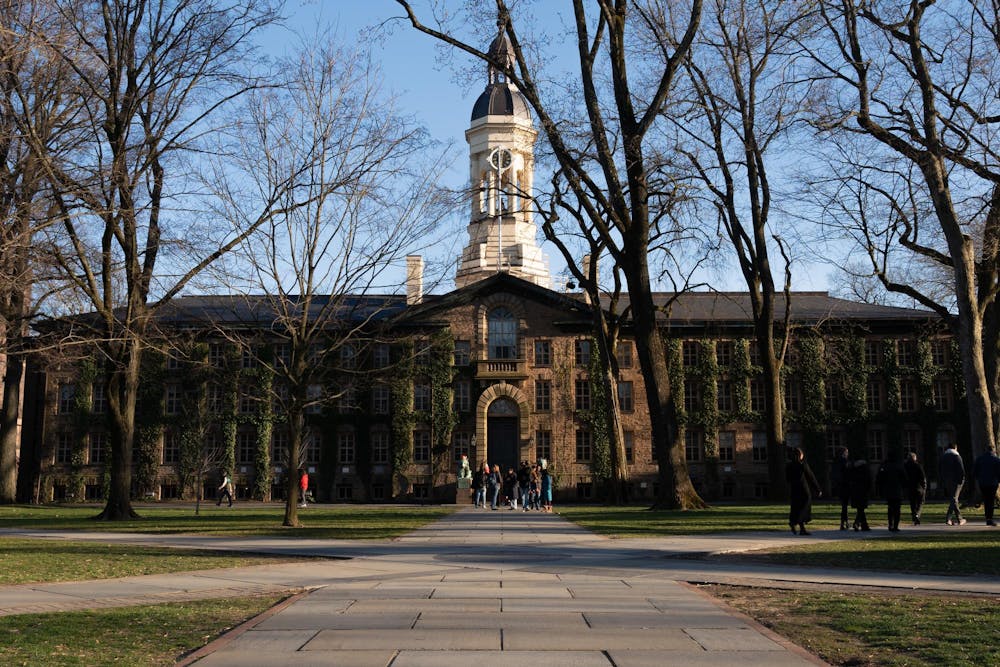 A large rectangular stone building with a bell tower is bathed in late afternoon light. Several groups stand around the lawn in front of the building. 