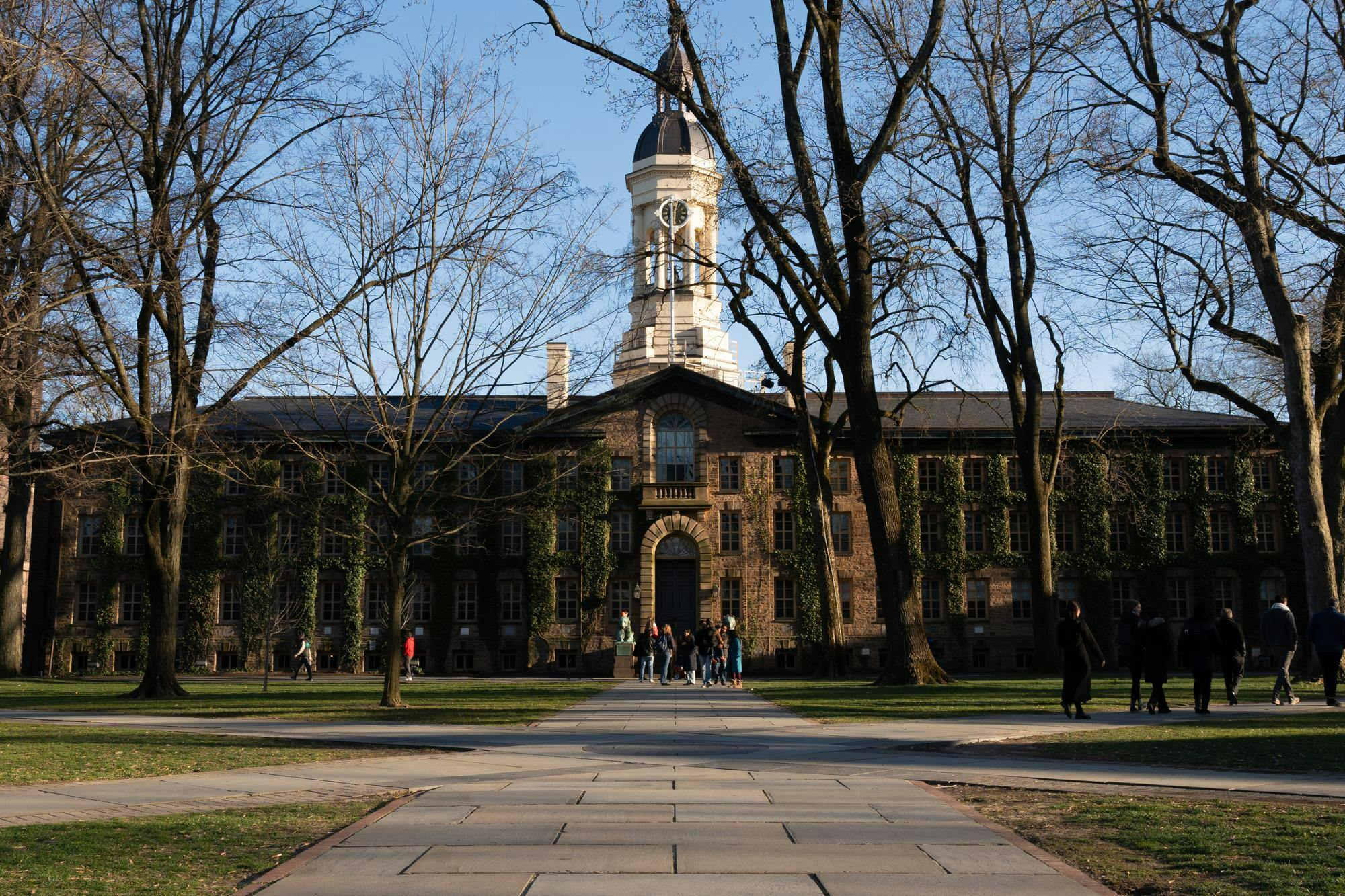 A large rectangular stone building with a bell tower is bathed in late afternoon light. Several groups stand around the lawn in front of the building. 