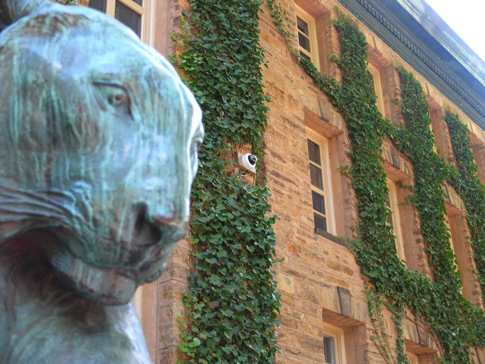 A security camera is mounted on an ivy clad stone building. A bronze tiger statue is visible in the foreground.