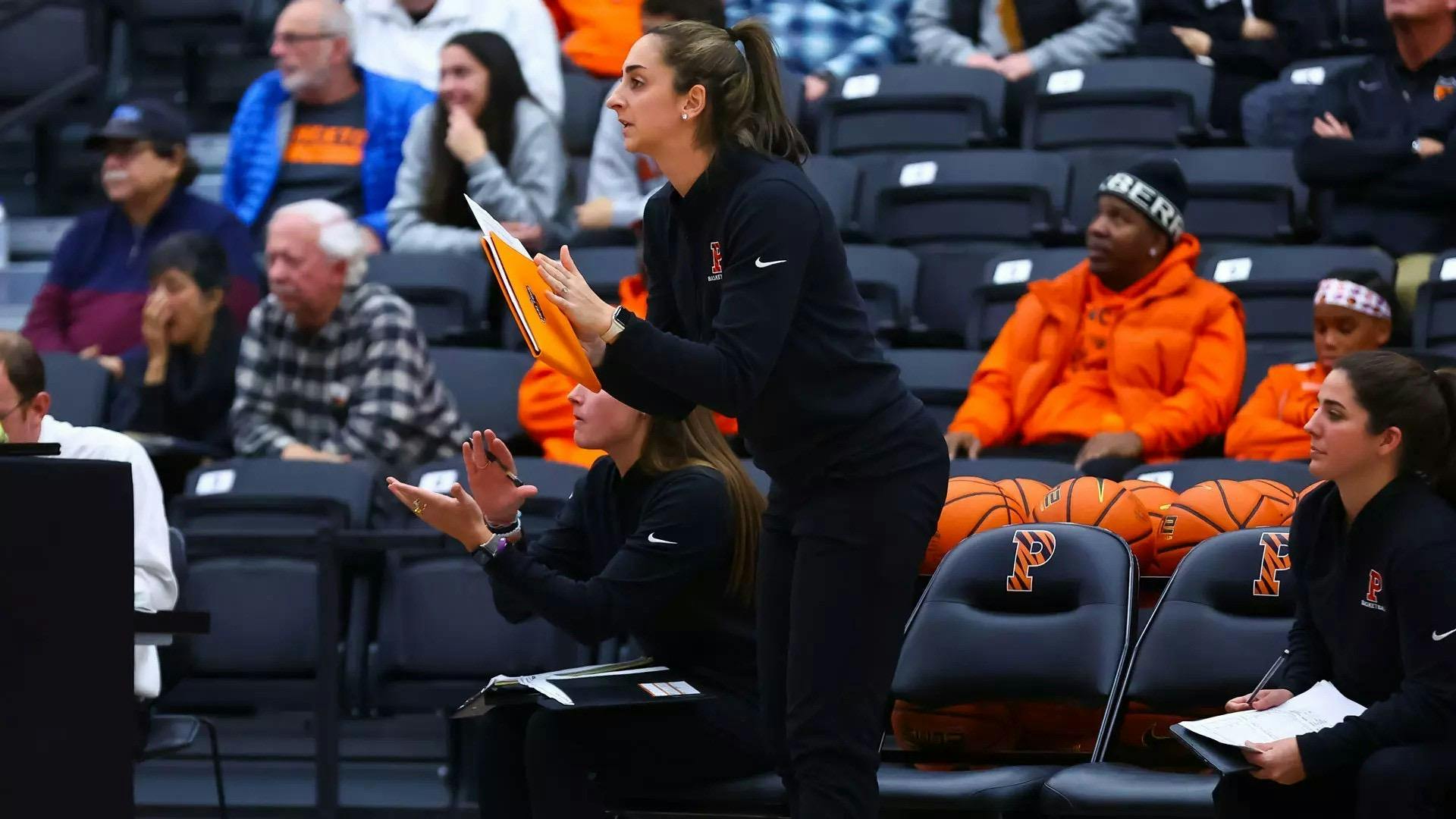 A women’s basketball coach stands on the sideline holding a clipboard during a game at Jadwin Gymnasium.