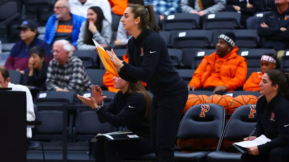A women’s basketball coach stands on the sideline holding a clipboard during a game at Jadwin Gymnasium.