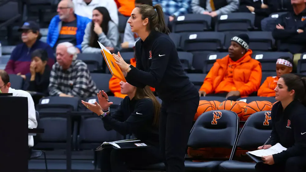 A women’s basketball coach stands on the sideline holding a clipboard during a game at Jadwin Gymnasium.