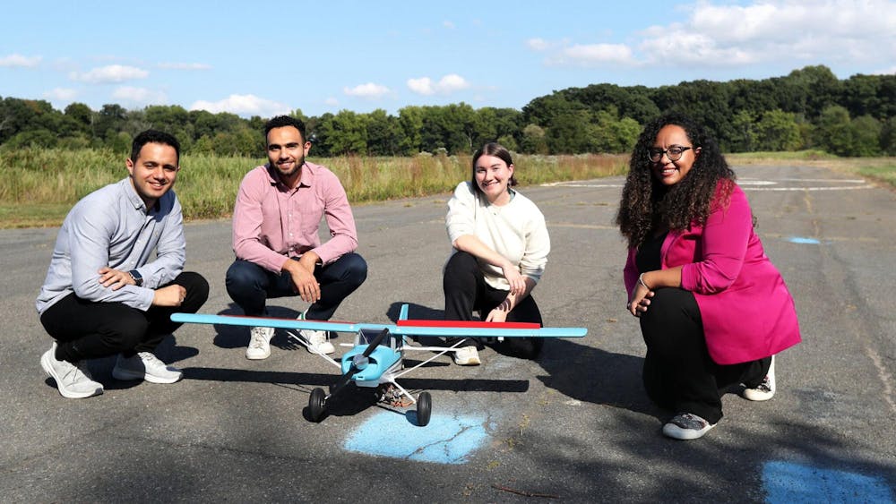 Four people are crouching outside on an asphalt surface. In the foreground, there is a small blue and red model plane. The background contains a blue sky and green trees and bushes.