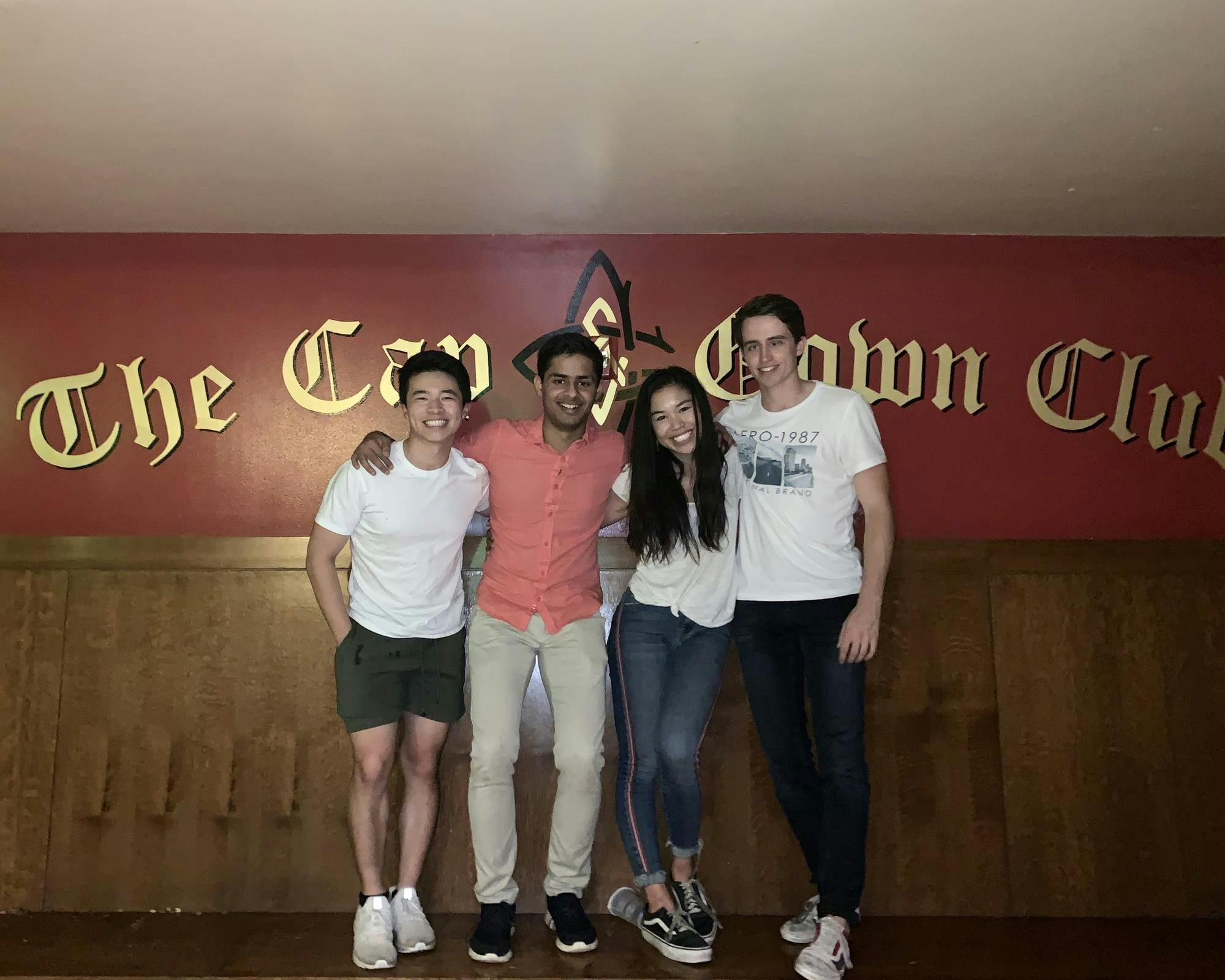 Four people stand with their arms around each other on a bench in front of a red wall that reads 'The Cap & Gown Club'