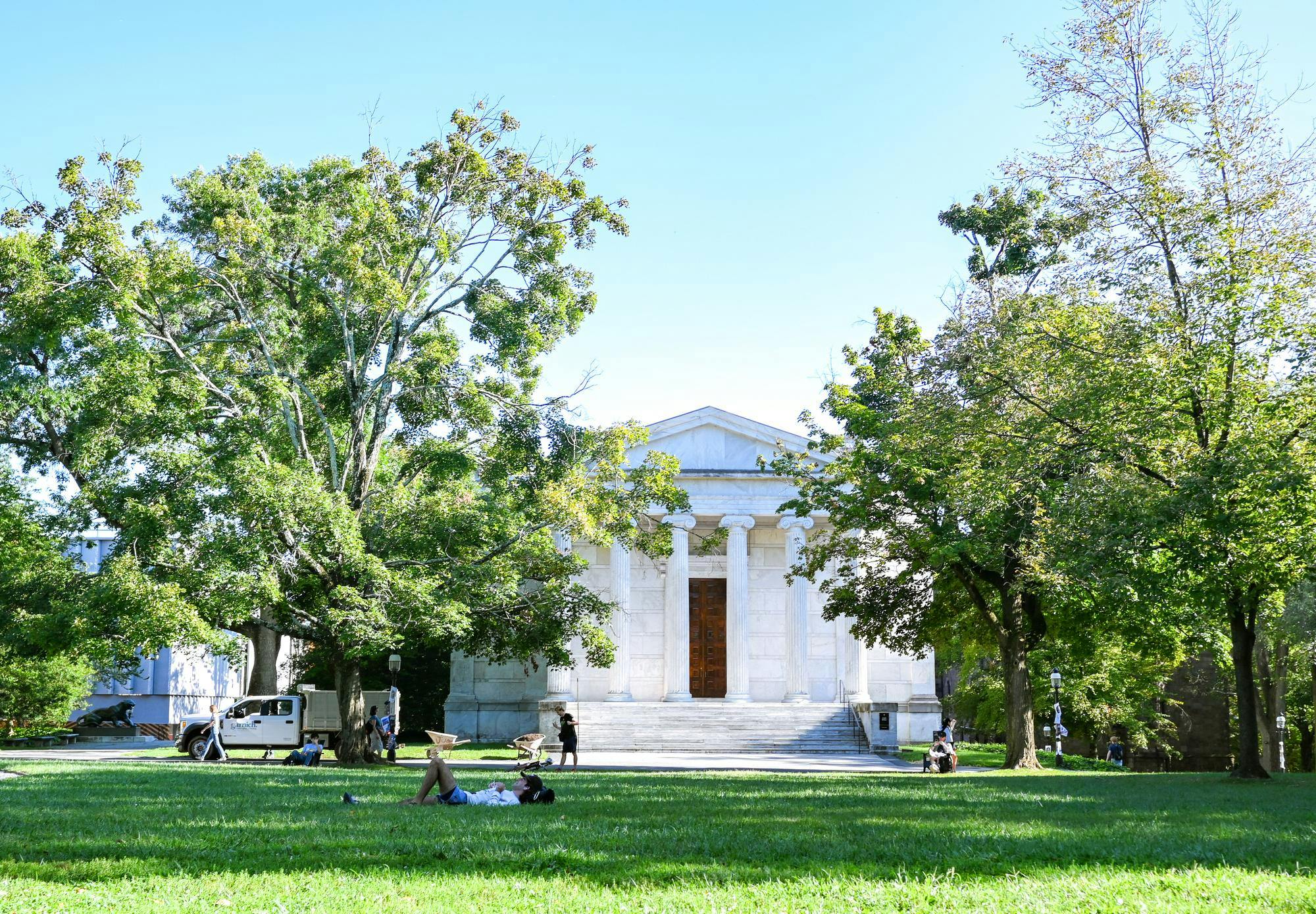 A building with ionic columns flanked by trees overlooks a grassy field. The sun shines overhead.