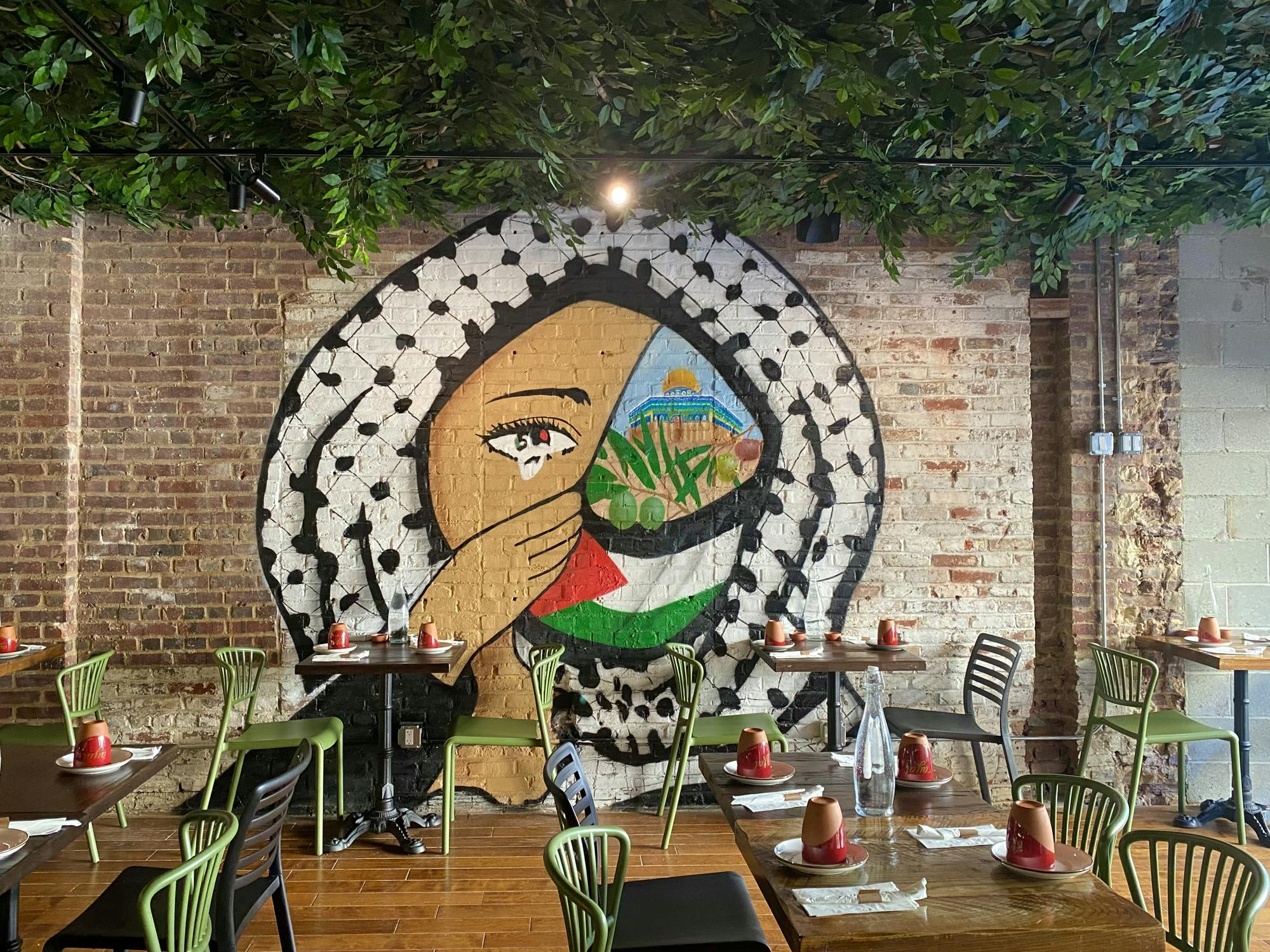 An interior of a restaurant with tables, green chairs, a brick wall with a mural, and a forest-lined ceiling.