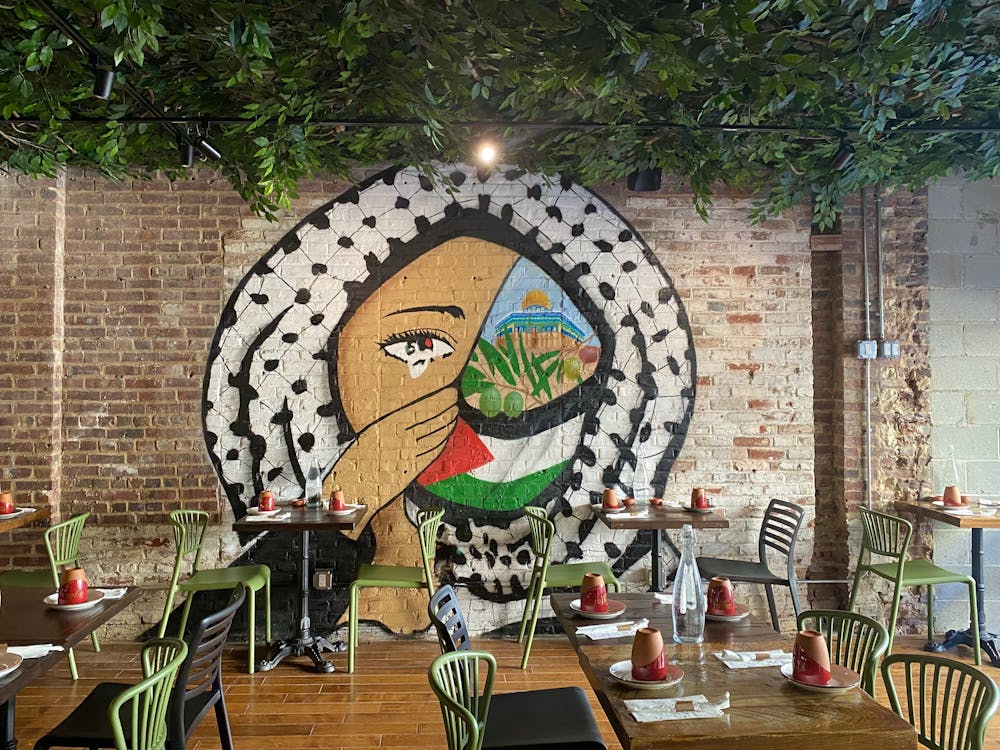 An interior of a restaurant with tables, green chairs, a brick wall with a mural, and a forest-lined ceiling.