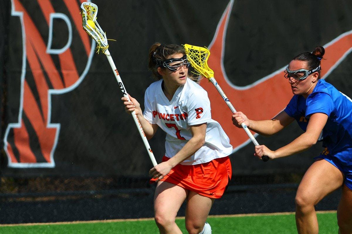 A woman in an orange and white uniform cradles the lacrosse ball.