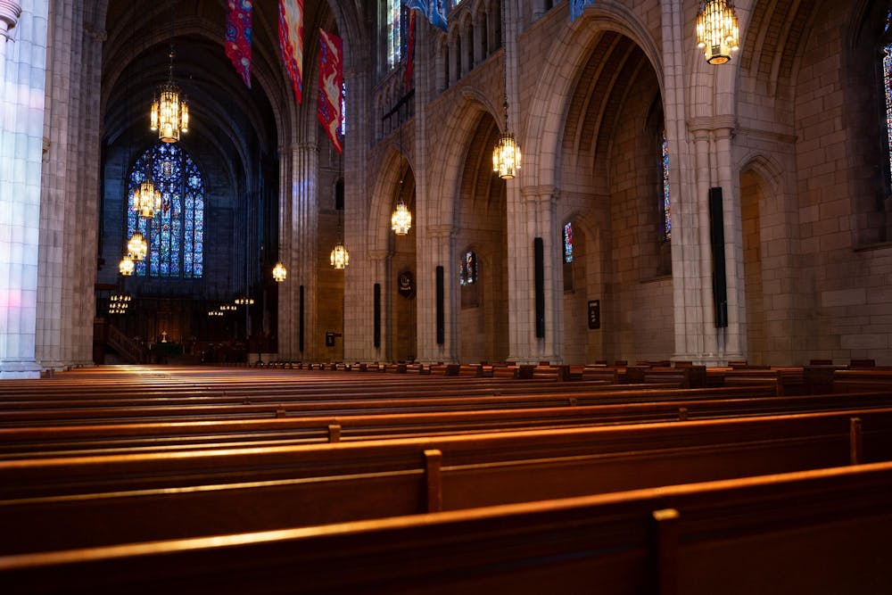 Brightly lit interior of a cavernous room filled with wooden pews and chandeliers hang overhead. 
