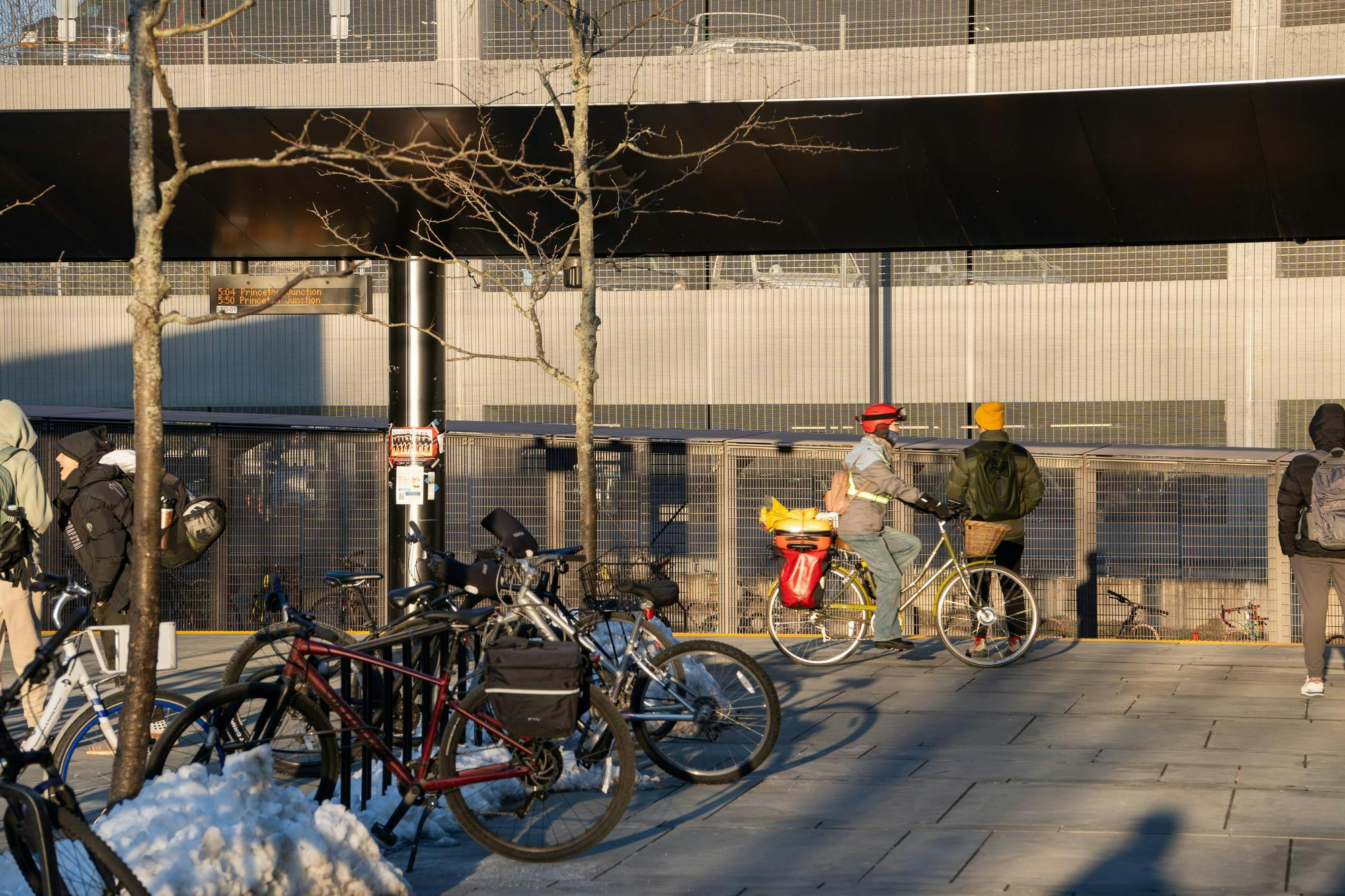At the Dinky train station, people wait for train by the tracks. Bikes are in the foreground locked to the bike stand. The sun is shining and shadows are seen next to the bikes and people.