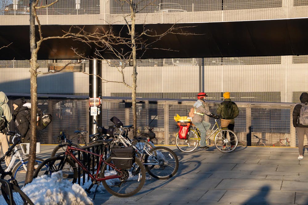 At the Dinky train station, people wait for train by the tracks. Bikes are in the foreground locked to the bike stand. The sun is shining and shadows are seen next to the bikes and people.