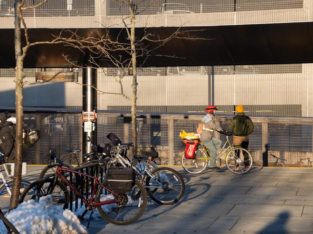 At the Dinky train station, people wait for train by the tracks. Bikes are in the foreground locked to the bike stand. The sun is shining and shadows are seen next to the bikes and people.