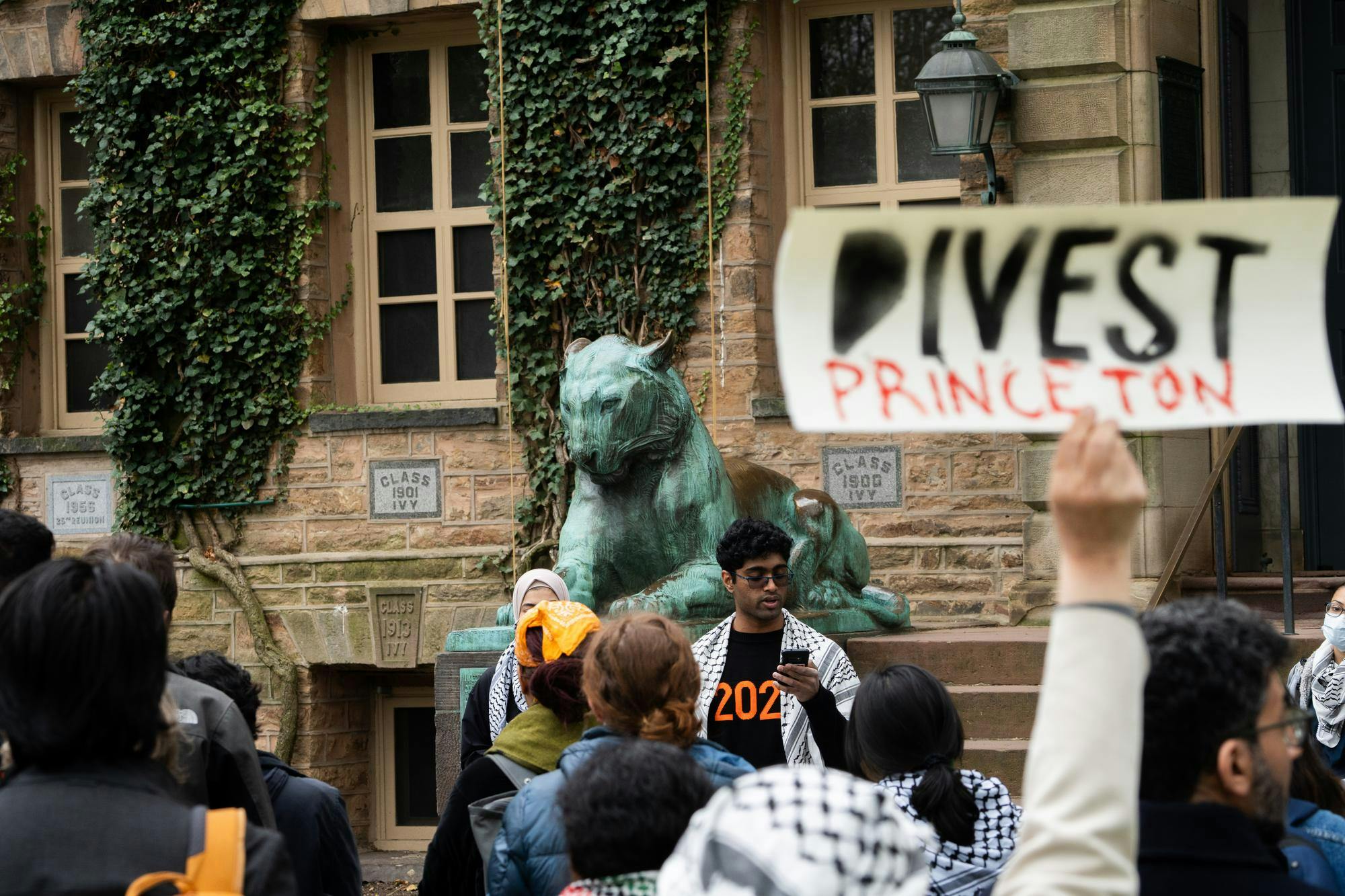 A man wearing dark glasses and a white keffiyeh over a black and orange sweater that reads “2026” gives a speech in front of the steps of an ivy-covered building. In the foreground a sign reads “DIVEST PRINCETON.”