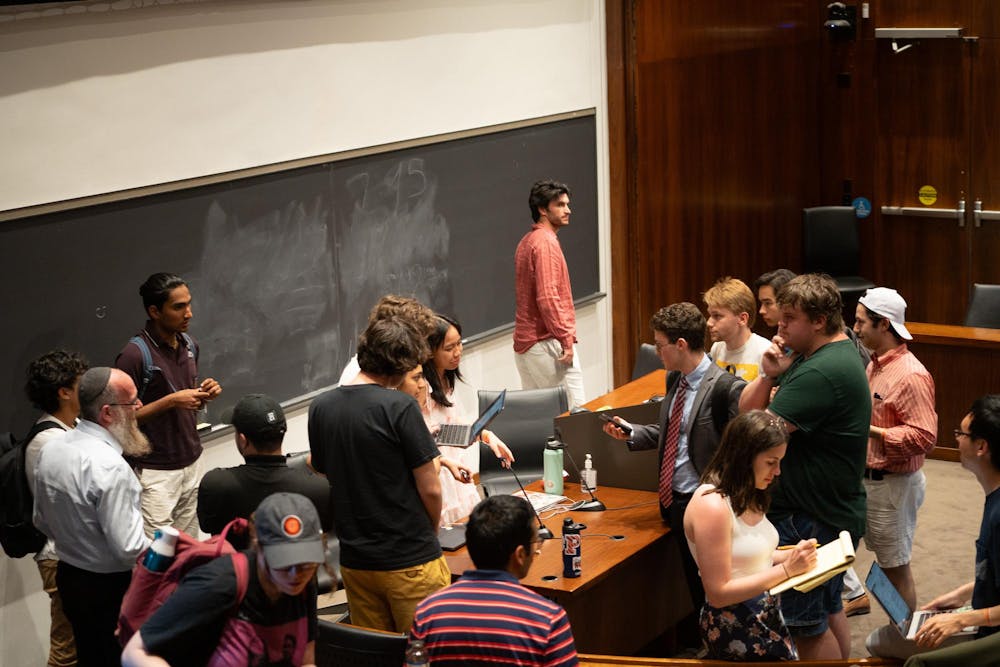 Room with wood paneling and a large chalkboard, shot from a high angle. Students mill around the forum in conversation.