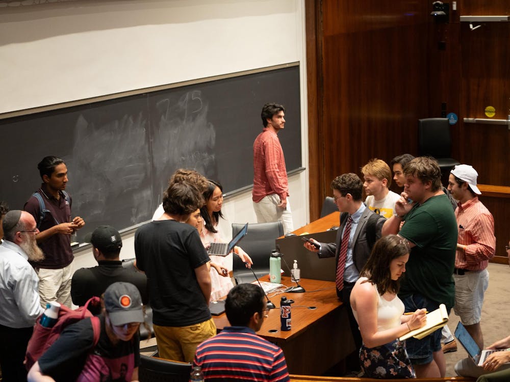 Room with wood paneling and a large chalkboard, shot from a high angle. Students mill around the forum in conversation.