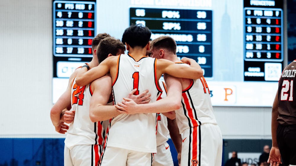 Several Princeton basketball players, wearing orange and white, huddle at the center of the court. In the background, there is a scoreboard and blue/light blue wall, and to the right is a Brown player wearing a brown and red jersey.