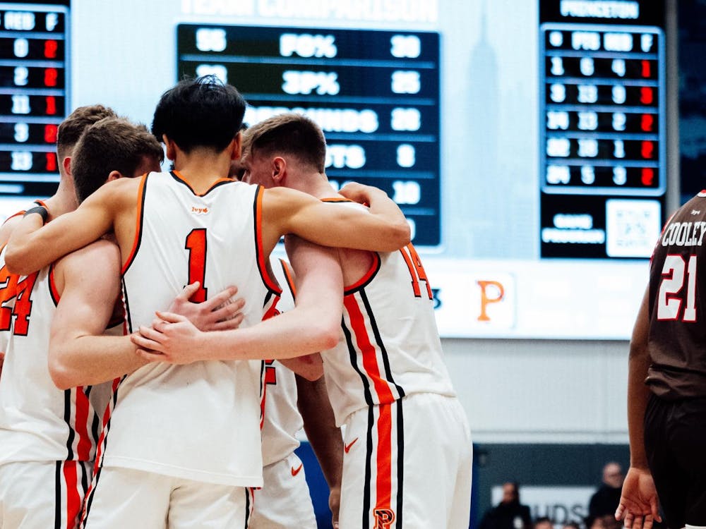 Several Princeton basketball players, wearing orange and white, huddle at the center of the court. In the background, there is a scoreboard and blue/light blue wall, and to the right is a Brown player wearing a brown and red jersey.