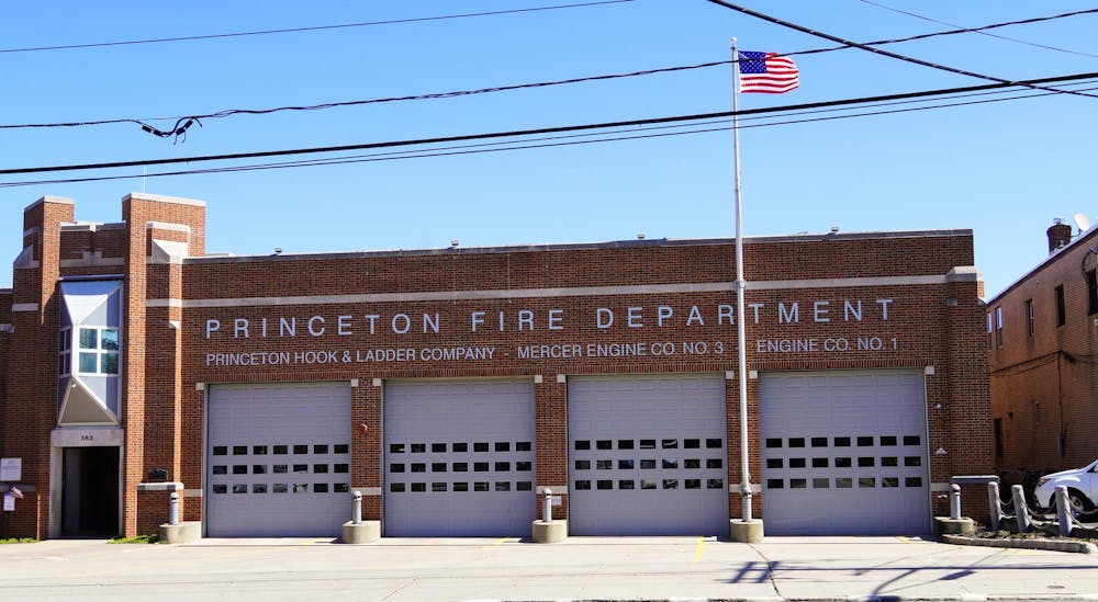 A wide, tan-brick building stands behind an American flag. Lettering on the building reads “Princeton Fire Department.”