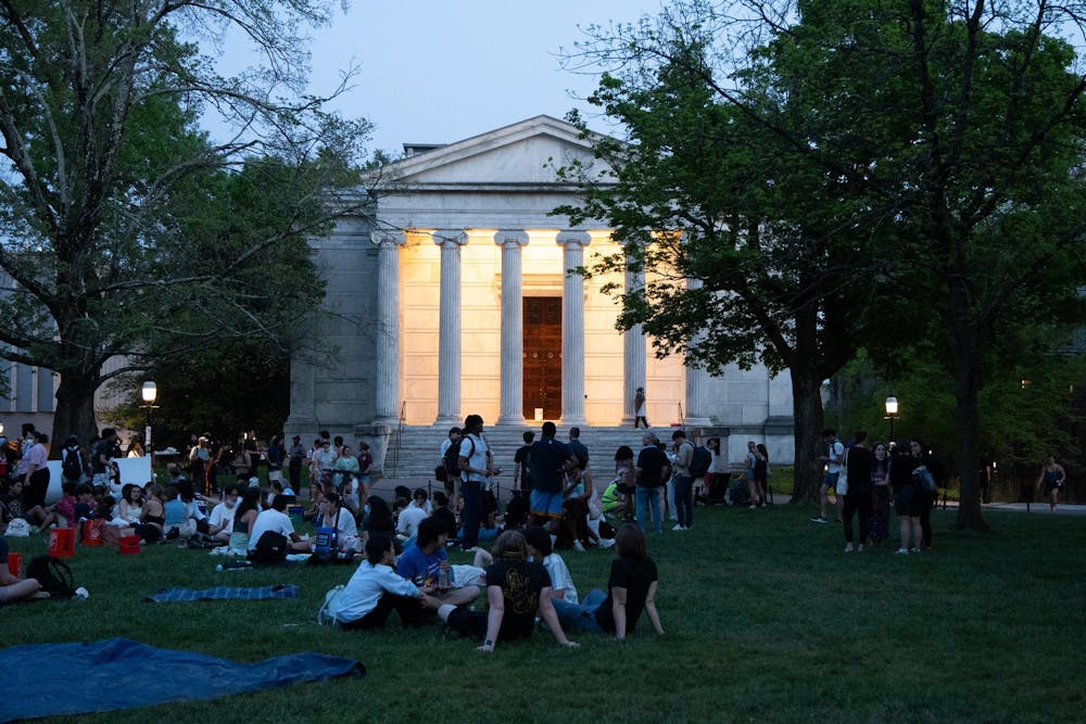 A building with columns sits behind several groups of people sitting on the ground. 