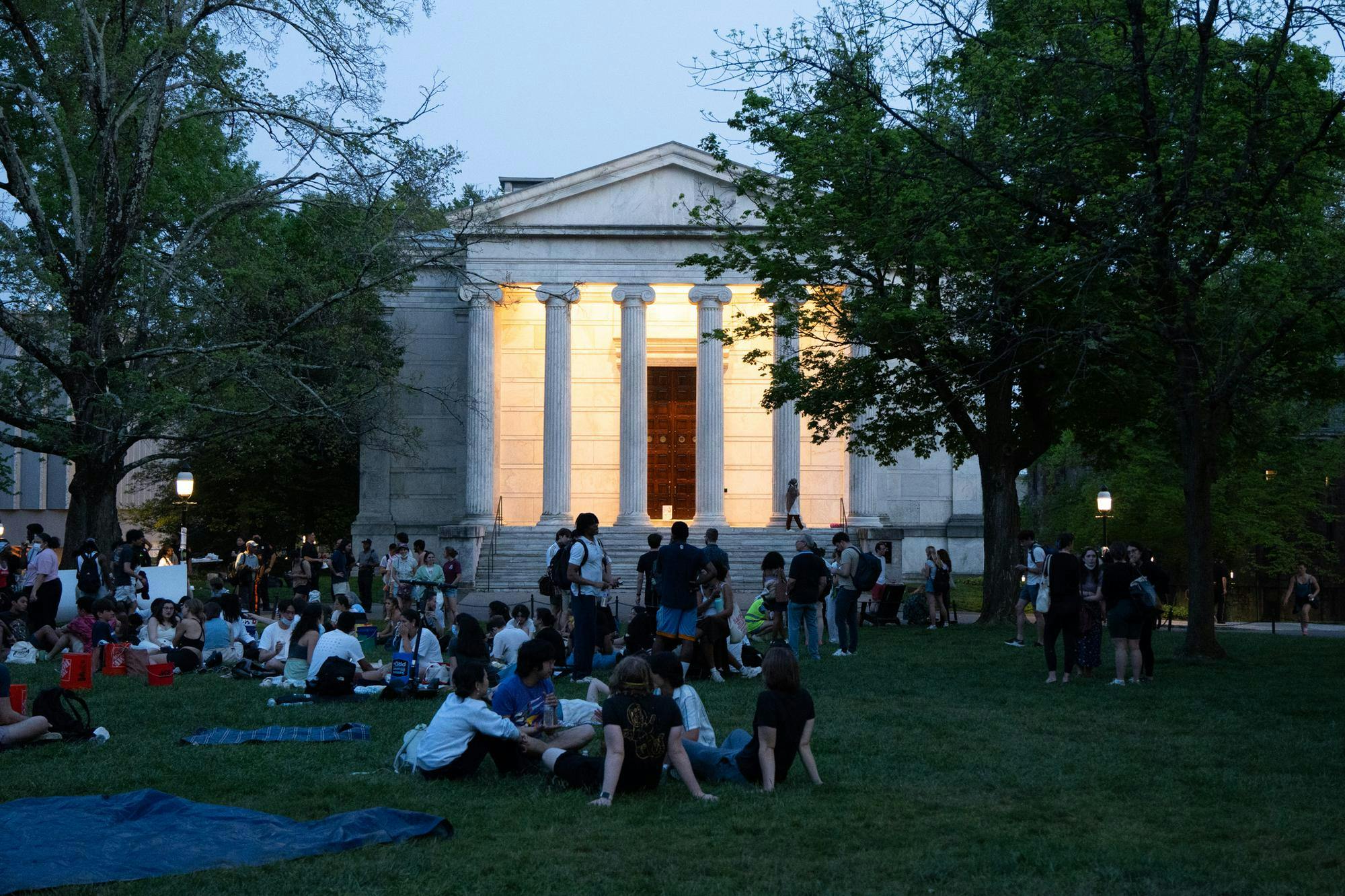 A building with columns sits behind several groups of people sitting on the ground. 