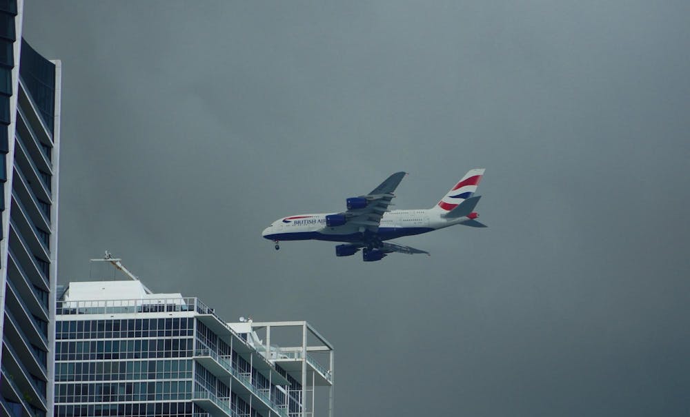 A British Airways Airbus A380 ascends over Miami, Florida following takeoff. The plane has a red and blue stripe and climbs over a cloudy sky and a blue glass-clad building.