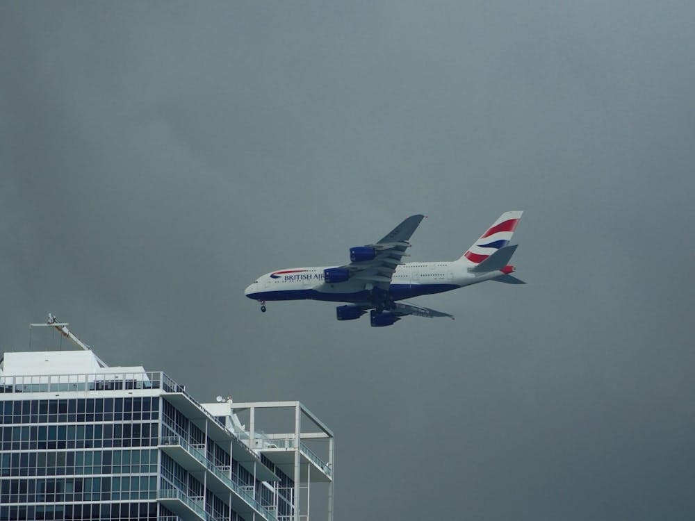 A British Airways Airbus A380 ascends over Miami, Florida following takeoff. The plane has a red and blue stripe and climbs over a cloudy sky and a blue glass-clad building.