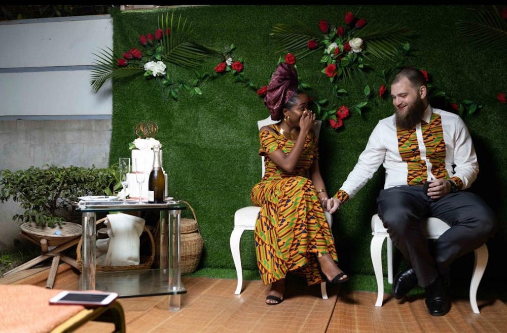 A man and woman are sitting on white chairs in front of a green background.