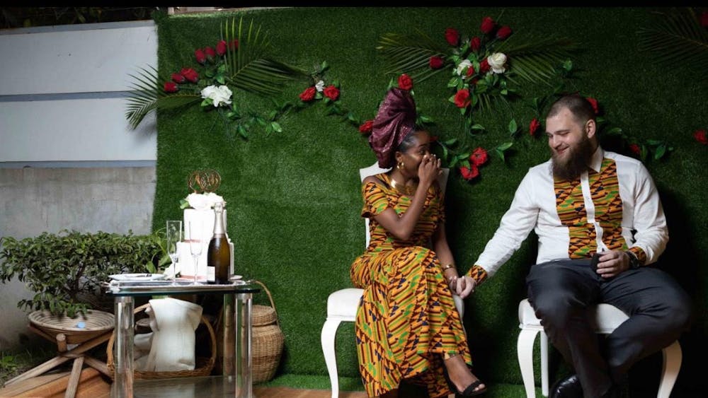 A man and woman are sitting on white chairs in front of a green background.