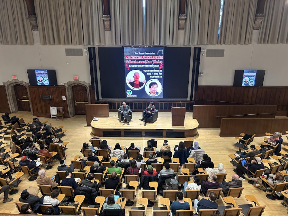 Two men sit in chairs facing an audience in a large lecture room. The man on the left is speaking. Behind them is a screen, showing a poster with the words "2nd Annual Conversation: Norman Finkelstein and Professor Max Weiss: A Conversation on Gaza."