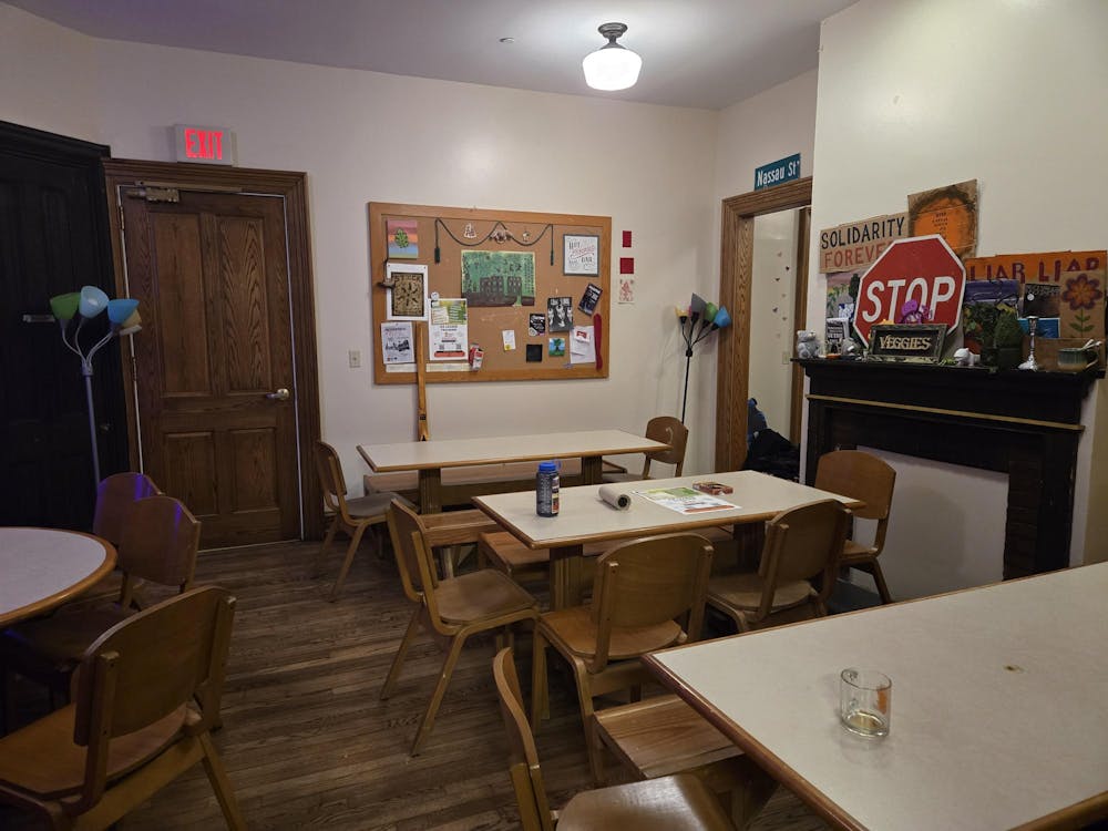 A small dining area with tables in the room. Wooden chairs are placed around the tables. A bulletin board rests on the back wall with several items attached to it. 