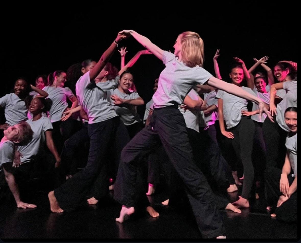 A group of dancers on stage with a black background. Dancers are wearing black pants and grey shirts.