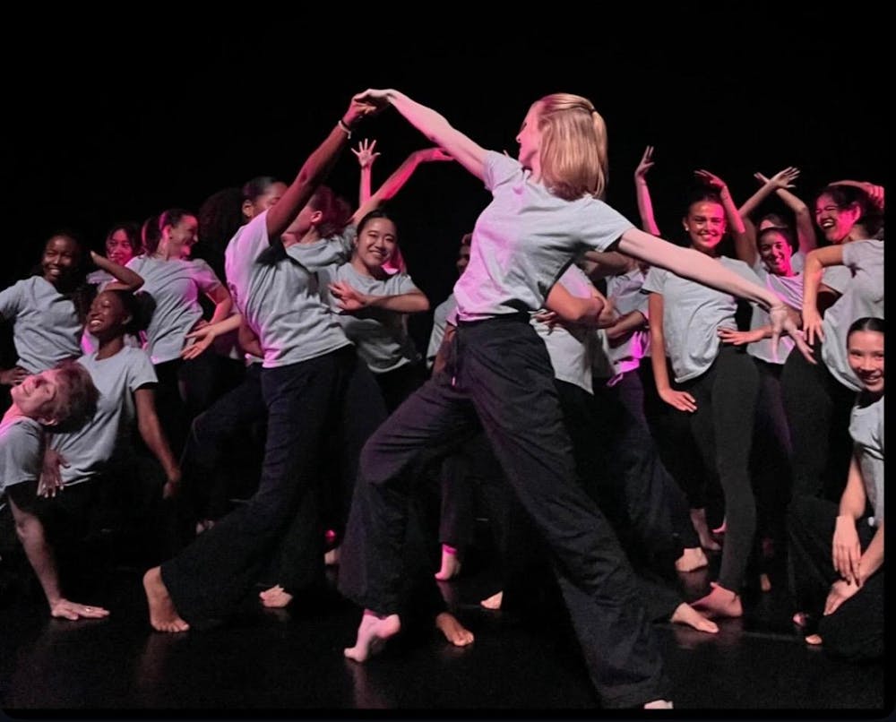 A group of dancers on stage with a black background. Dancers are wearing black pants and grey shirts.