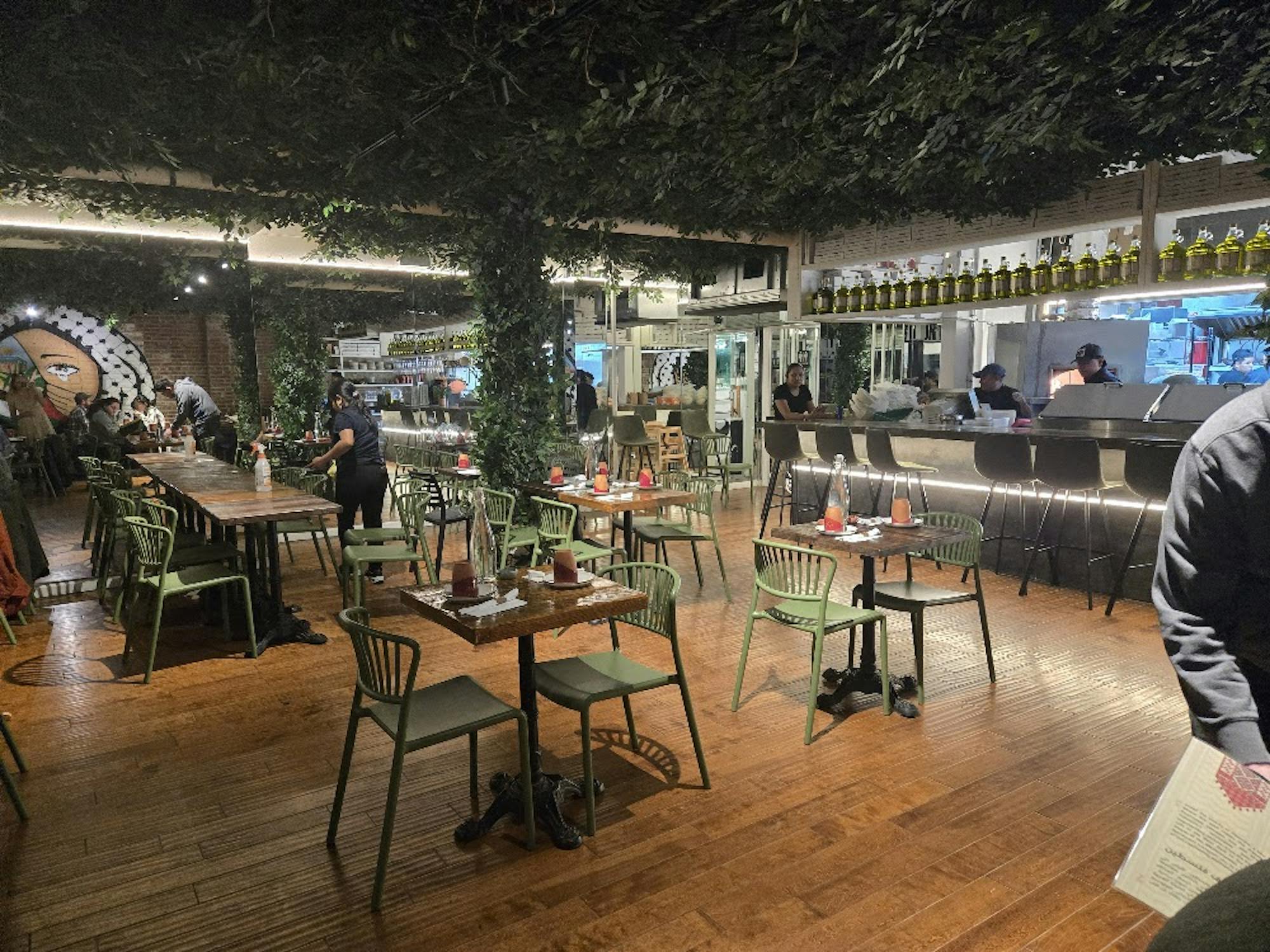 The interior design and layout of the restaurant, containing green chairs, wooden tables, and a plant-covered ceiling.