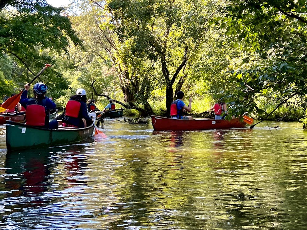 A group of canoes on the river filled with young people in life vests. Green trees fill the background.