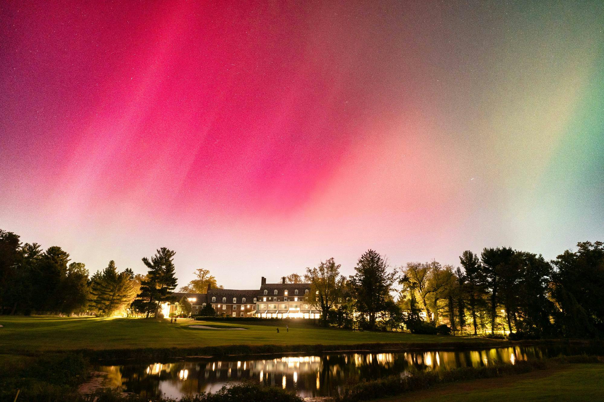 Streaks of pink, white, and green color the sky above a lake. Lights from Forbes Inn shine. 