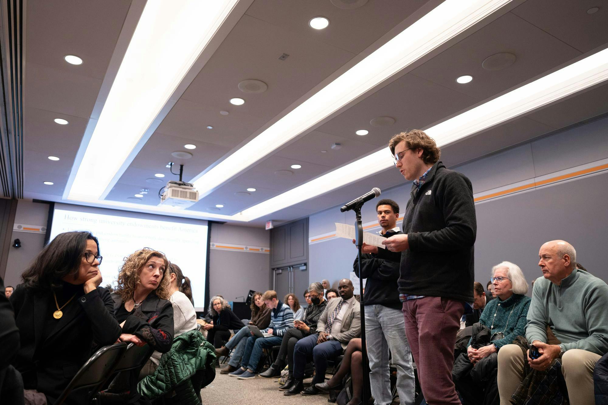 two seated women look up at a man speaking into a microphone 