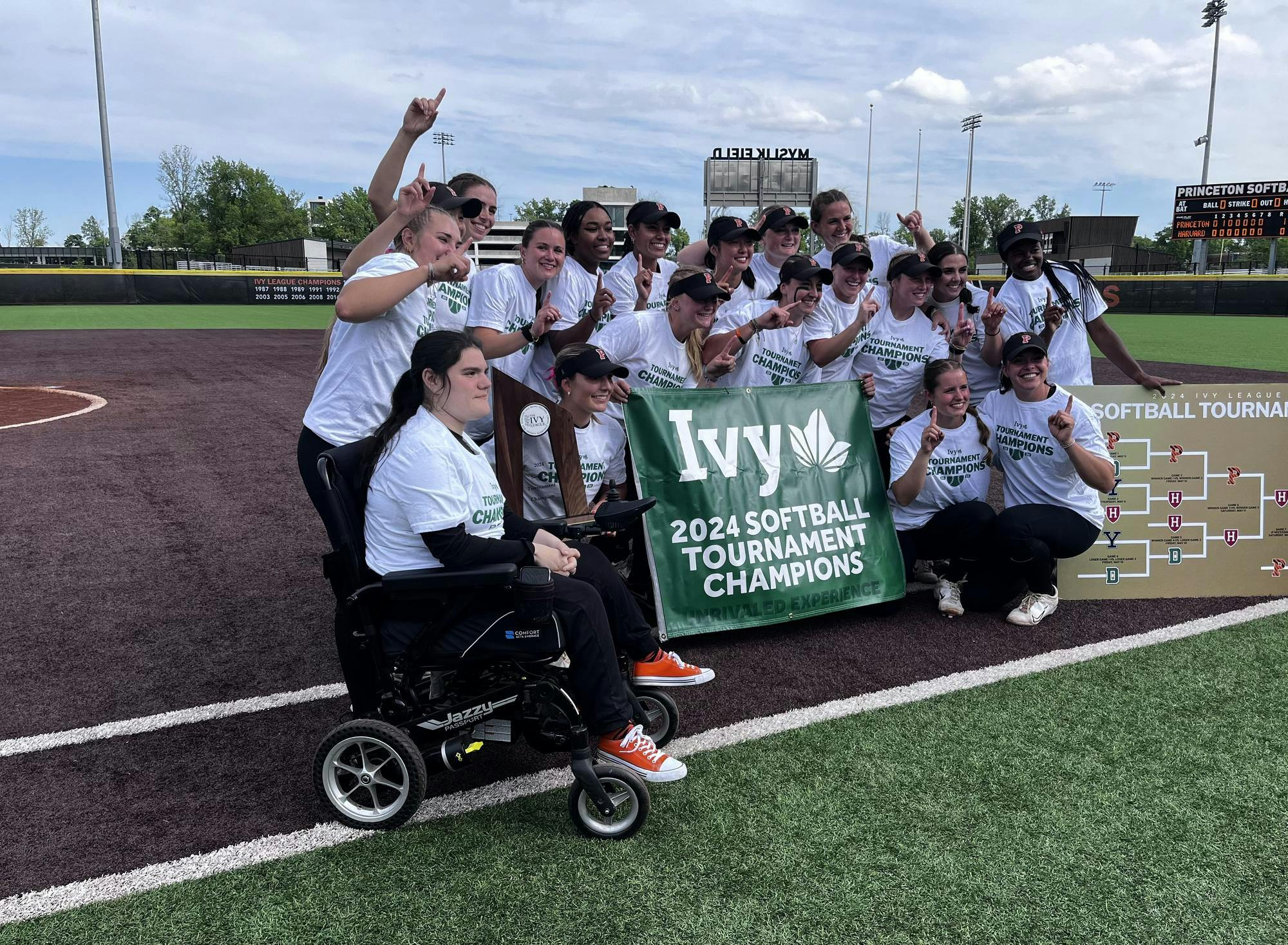 Group of about 16 softball players in white t-shirts that read "Ivy Tournament Champions" standing behind large green banner with the same wording.