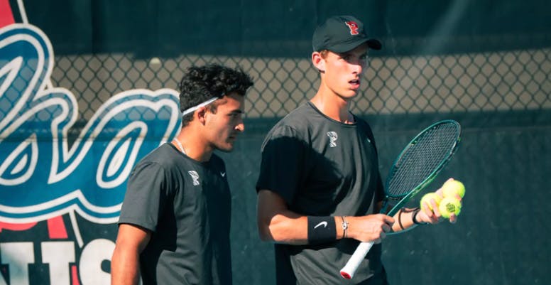 One man in a black uniform holding a racket and three tennis balls, talking to another man in a black uniform.
