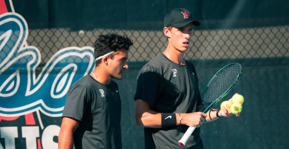 One man in a black uniform holding a racket and three tennis balls, talking to another man in a black uniform.