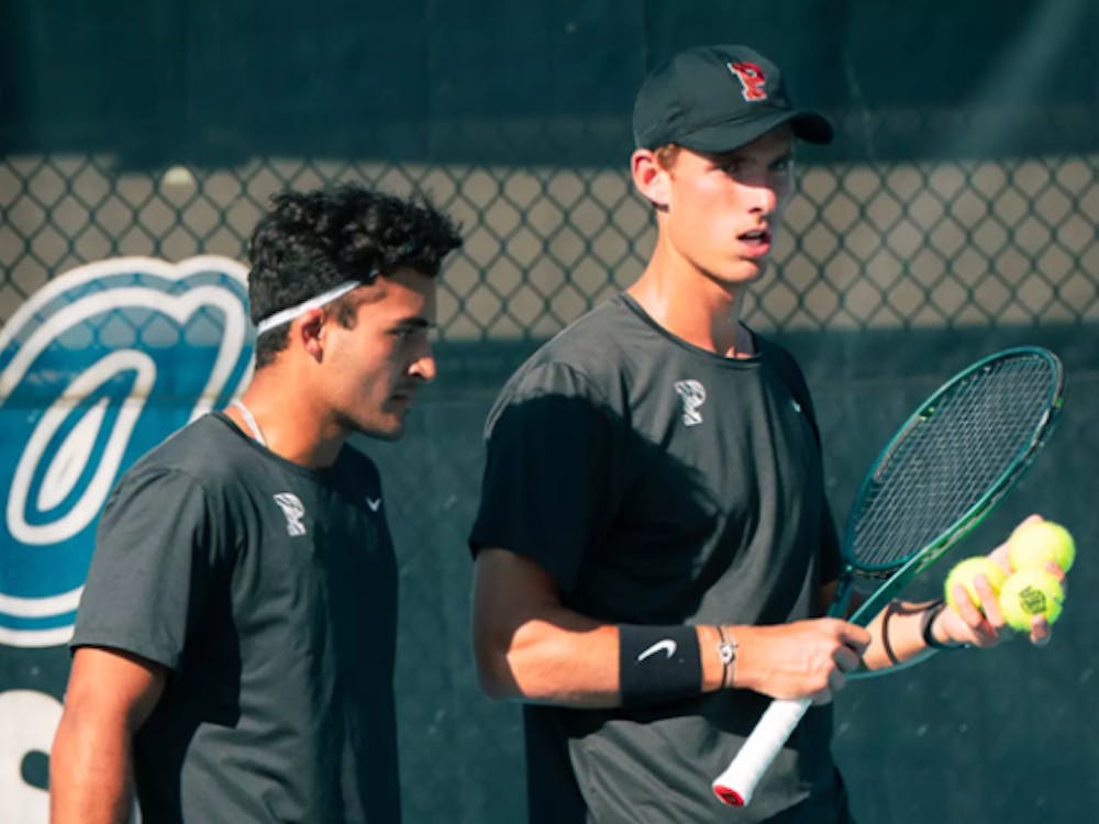 One man in a black uniform holding a racket and three tennis balls, talking to another man in a black uniform.