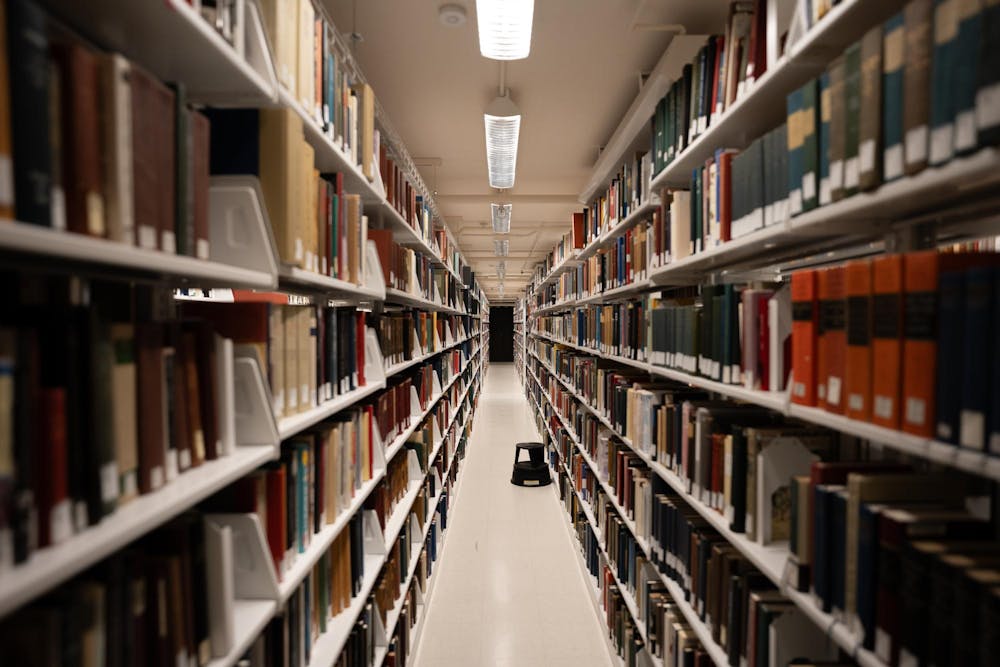 A long, blank, aisle with a white tile floor is the focus of the center of this photo, with a small black stool midway. Multicolored books in a blur line the sides.