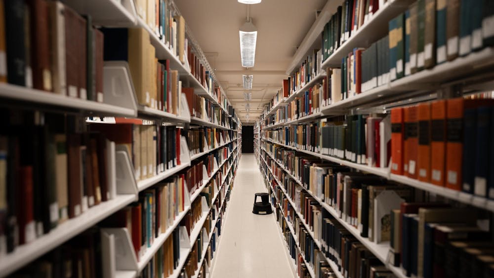 A long, blank, aisle with a white tile floor is the focus of the center of this photo, with a small black stool midway. Multicolored books in a blur line the sides.