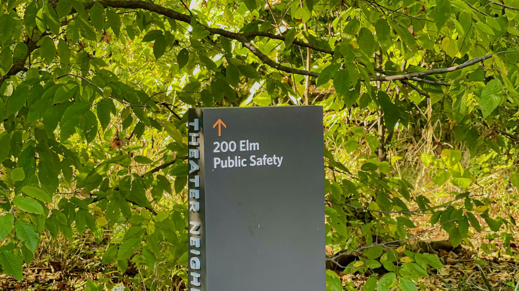 A sign surrounded by green foliage reads “200 Elm Public Safety” with an upward-pointing arrow.