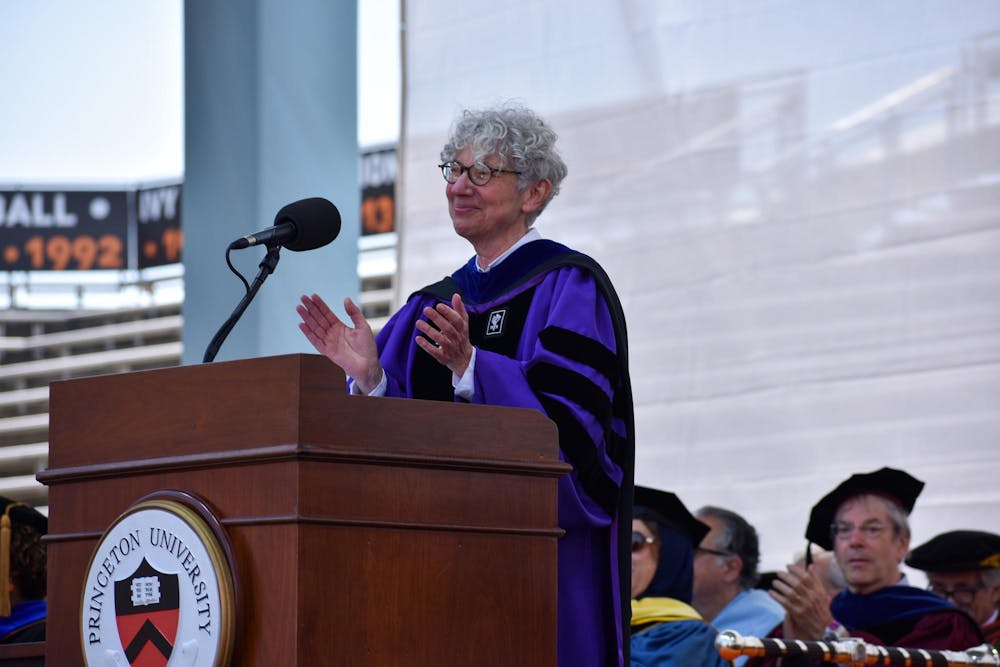 A woman in a purple graduation robe claps onstage behind a podium at a graduation ceremony. 