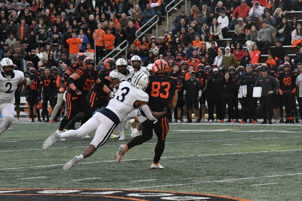 A football player wearing a black and orange jersey getting tackled by a player wearing a white and navy blue jersey as players, coaches, and fans watch it take place. 