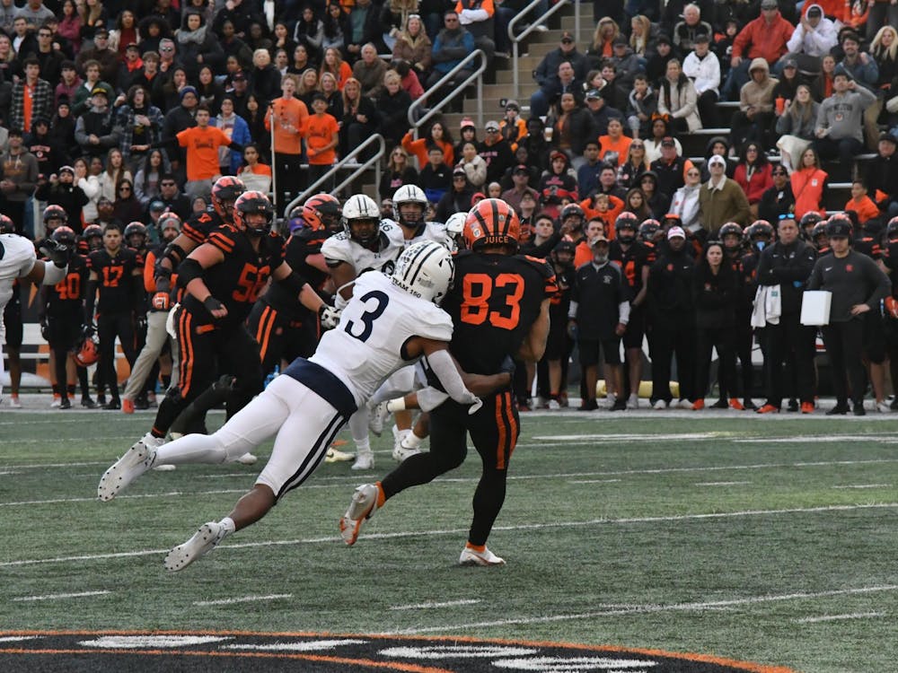 A football player wearing a black and orange jersey getting tackled by a player wearing a white and navy blue jersey as players, coaches, and fans watch it take place. 