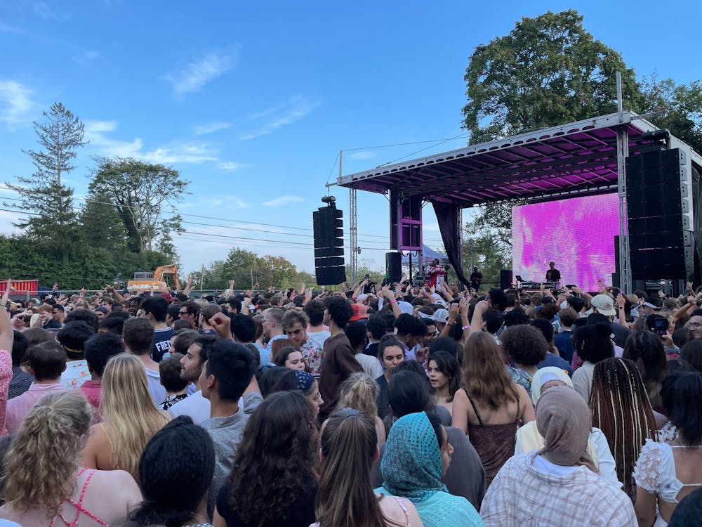 The backyard of Quadrangle club crowded with people facing a concert stage with large speakers suspended above the ground on either side. A$AP Ferg is performing on stage, and is wearing a white t-shirt and red hoodie. A DJ stands at a turntable behind him. A large screen displaying a pink and white tie-dye is directly behind the stage. Further behind the stage is a yellow construction vehicle. The area is surrounded by trees, and the sky is clear blue with some clouds.